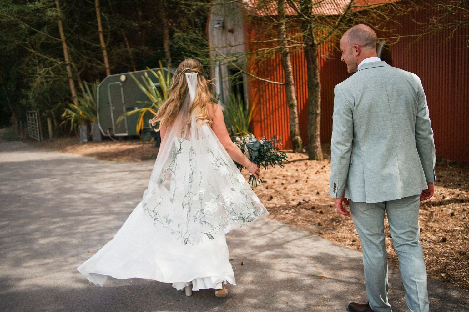 Bride in floral embroidered gown and groom in sage suit walk hand in hand outdoors.