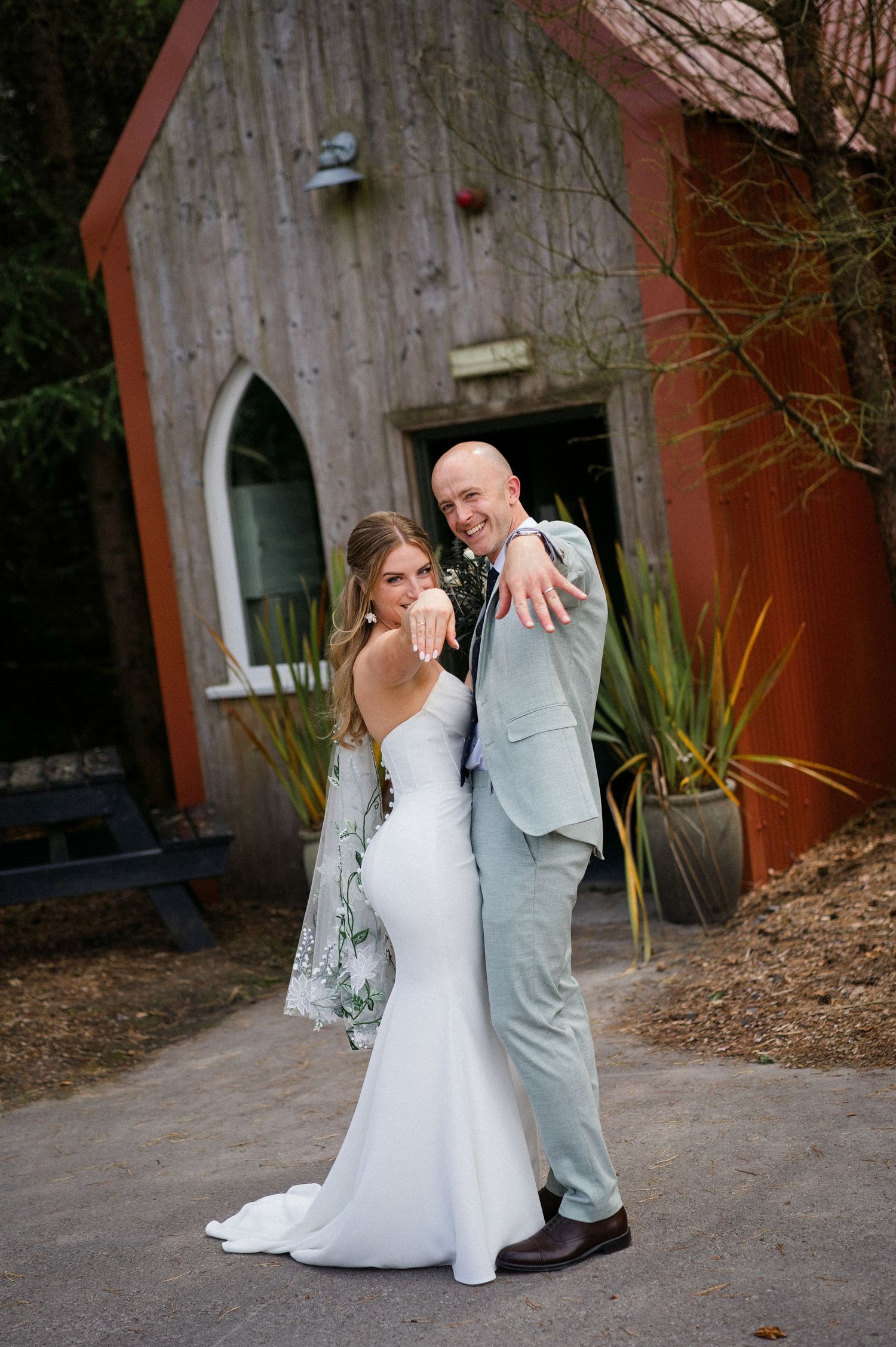 A bride and groom show off their wedding rings outside a rustic wooden chapel, smiling joyfully on their wedding day.