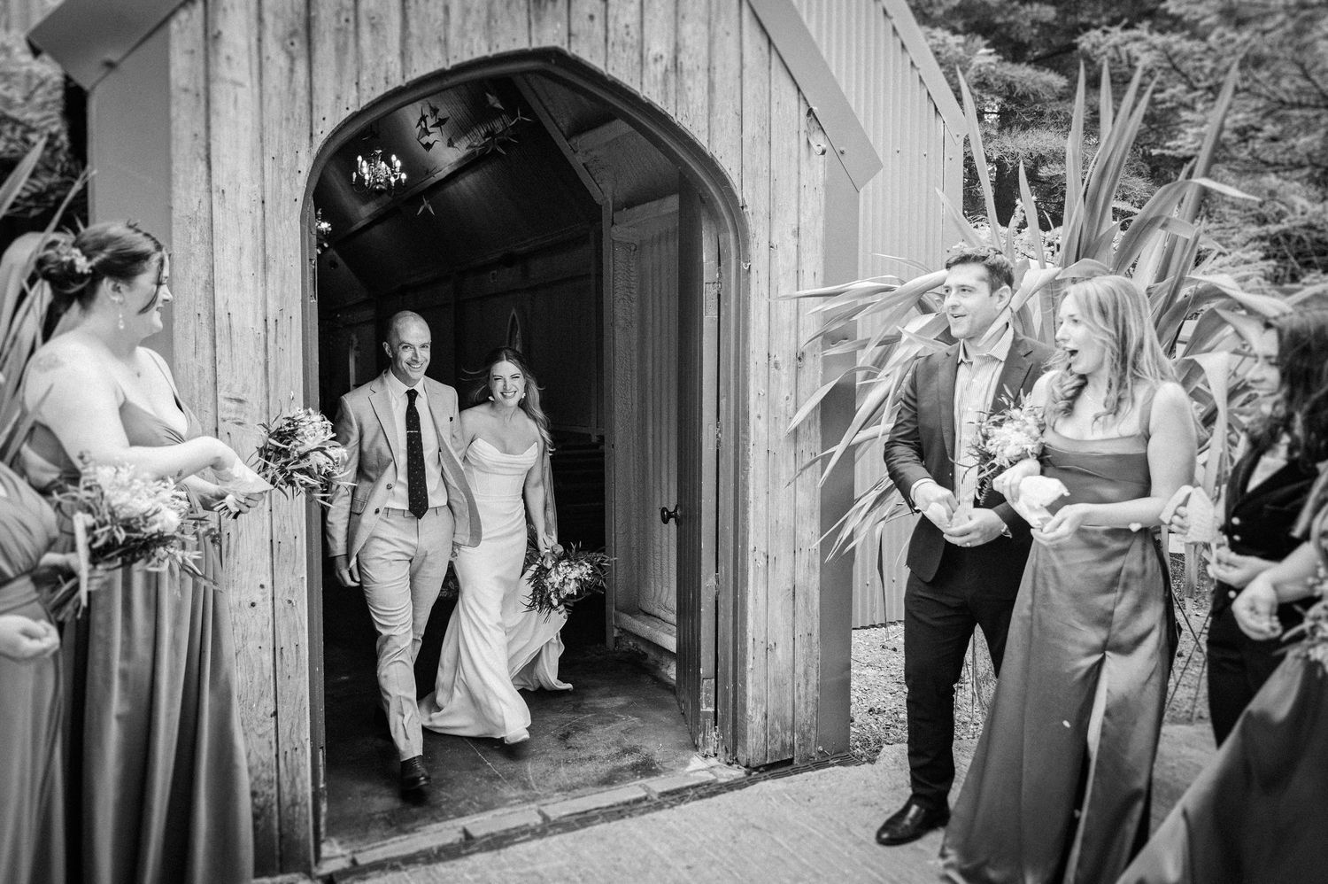 Bride and groom exiting rustic chapel doorway greeted by wedding party in black and white photo.