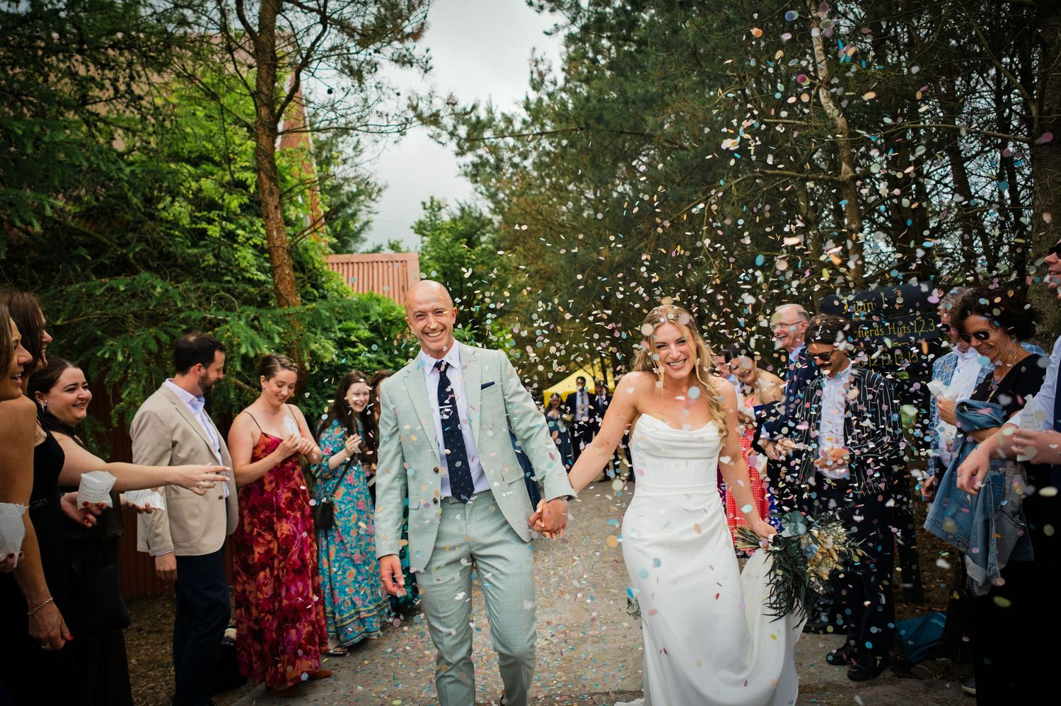 Joyful newlyweds walk through a colorful confetti shower surrounded by cheering wedding guests outdoors.