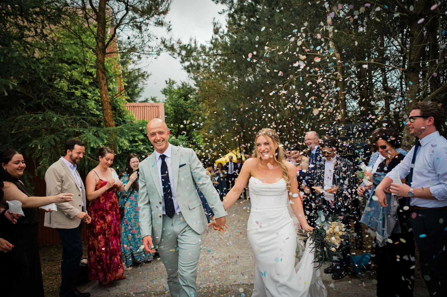 Newlywed couple walks hand in hand through a shower of colorful confetti surrounded by cheering wedding guests.