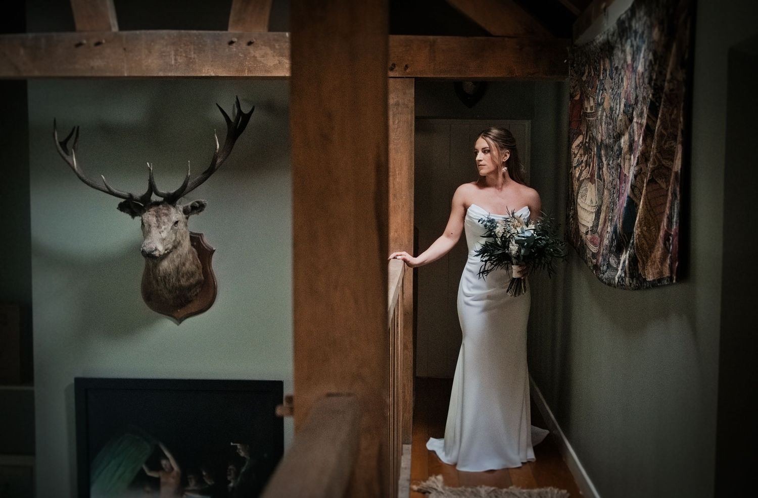 Elegant bride in fitted gown holds bouquet beside rustic wooden beam and deer mount.