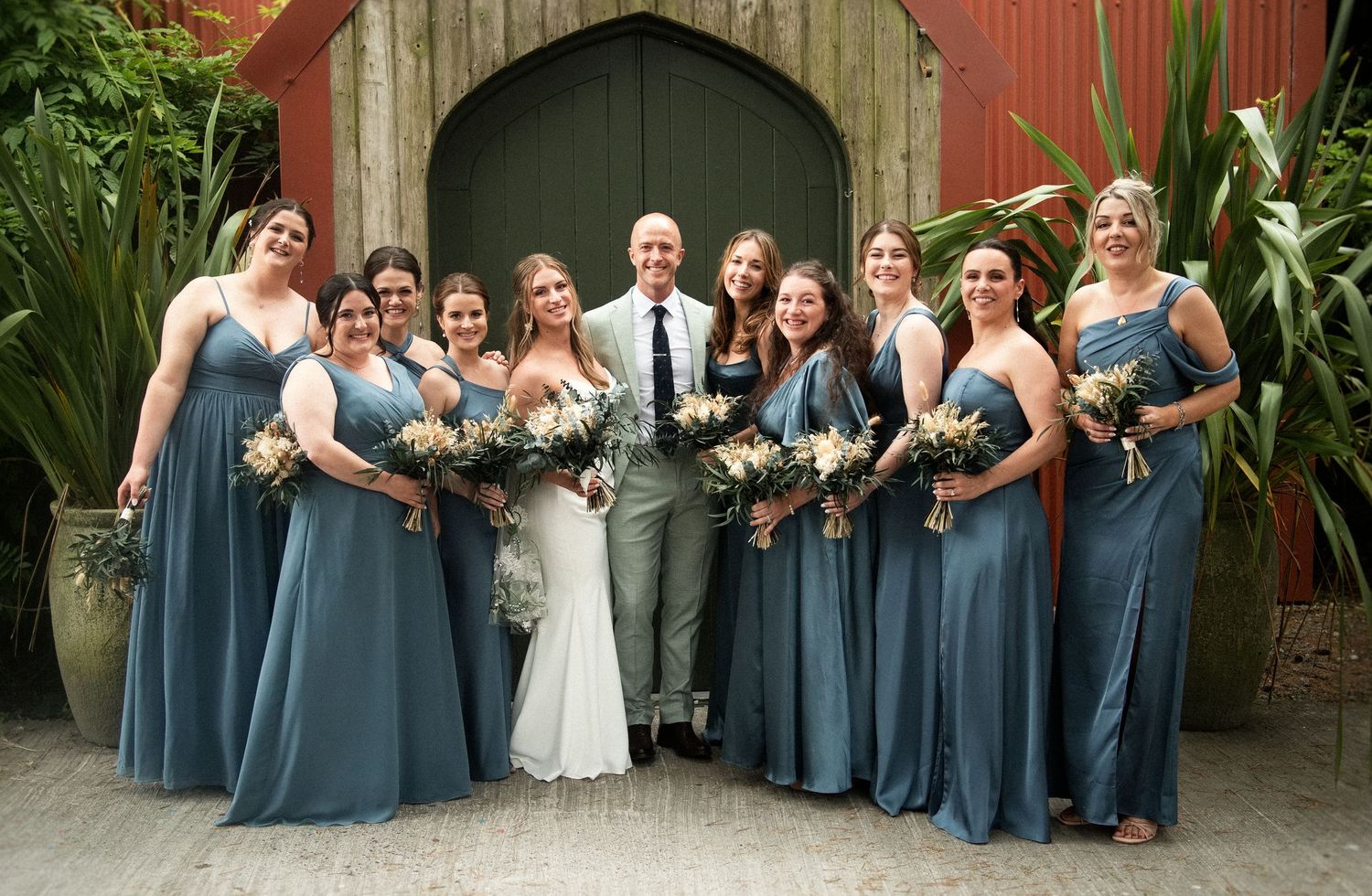 Groom poses with ten bridesmaids in dusty blue gowns holding bouquets outside a rustic barn venue.