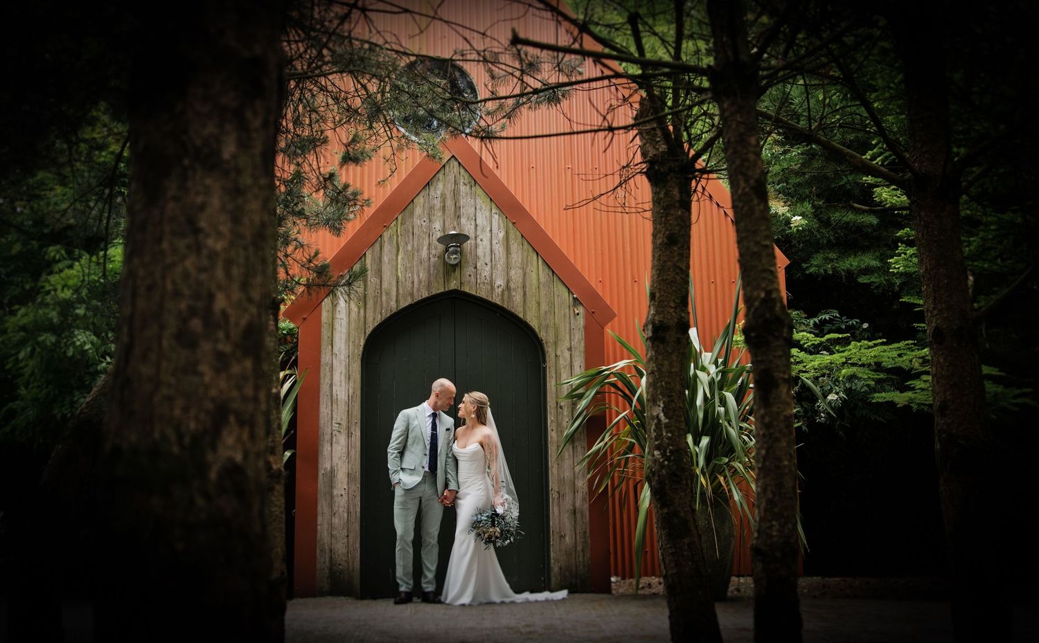 Bride and groom pose romantically before a rustic orange chapel surrounded by trees.