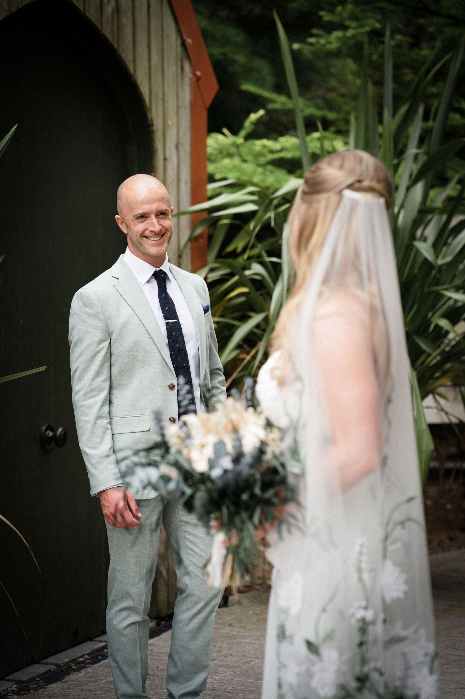 Groom in sage suit smiles warmly at bride during outdoor first look moment before wedding.