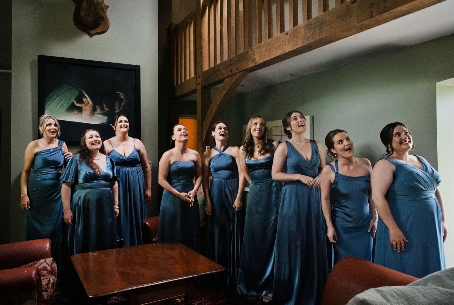 Eight bridesmaids in matching teal satin dresses standing together reacting with joy indoors.