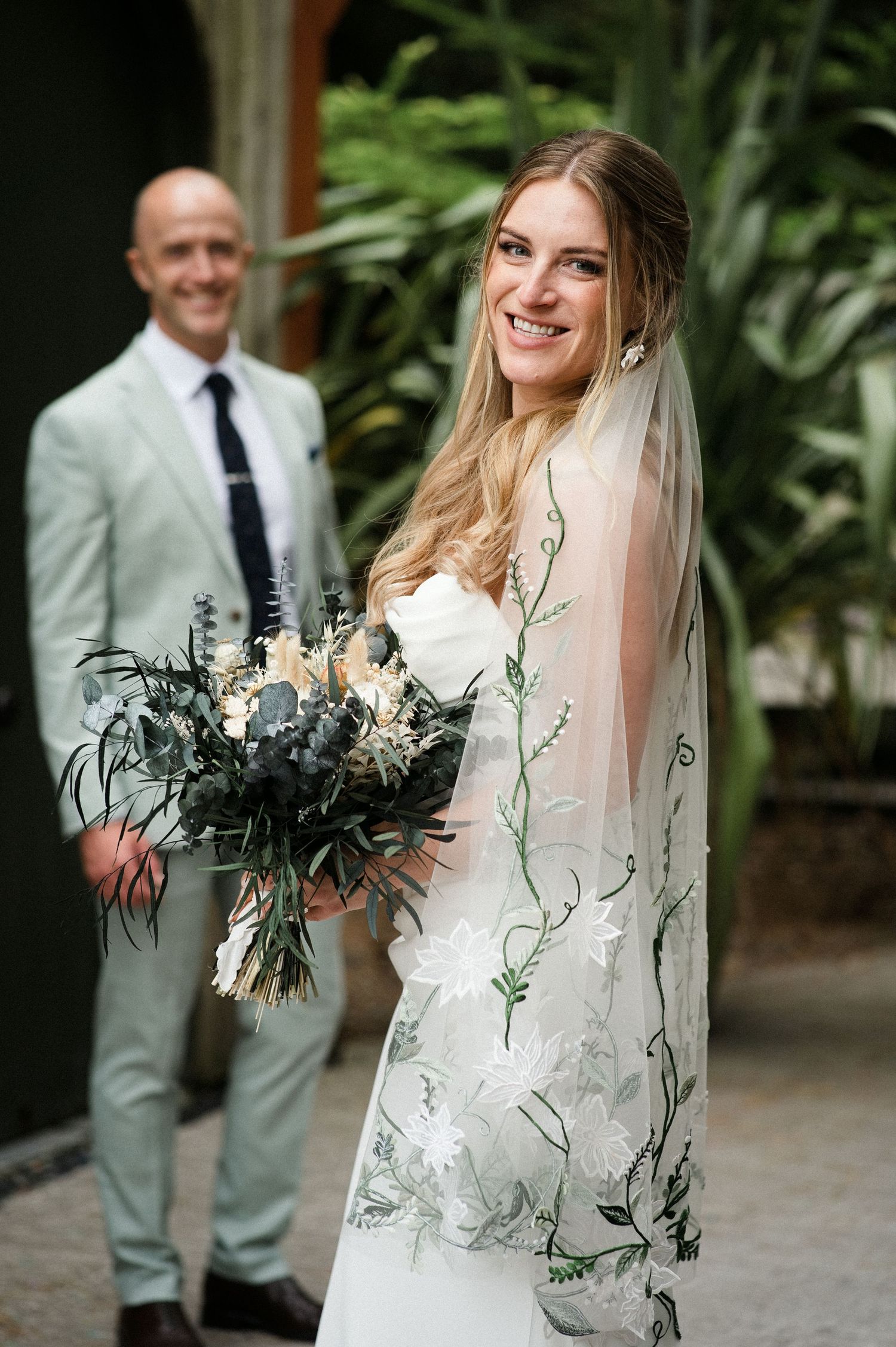 Smiling bride in white gown with floral veil holding bouquet, groom in mint suit standing behind her outdoors.