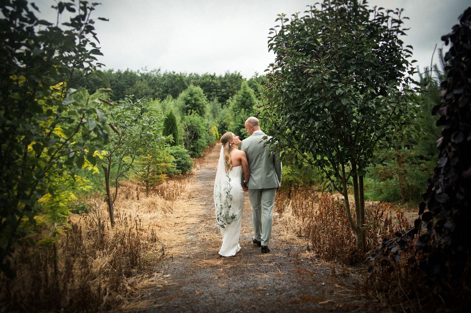 Bride and groom walking hand in hand through a tree-lined path.