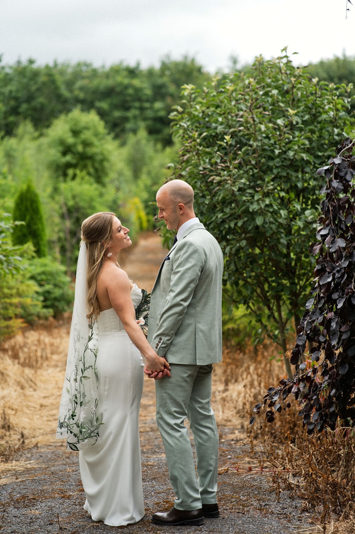 Bride and groom holding hands gazing at each other on a gravel path surrounded by lush green trees.