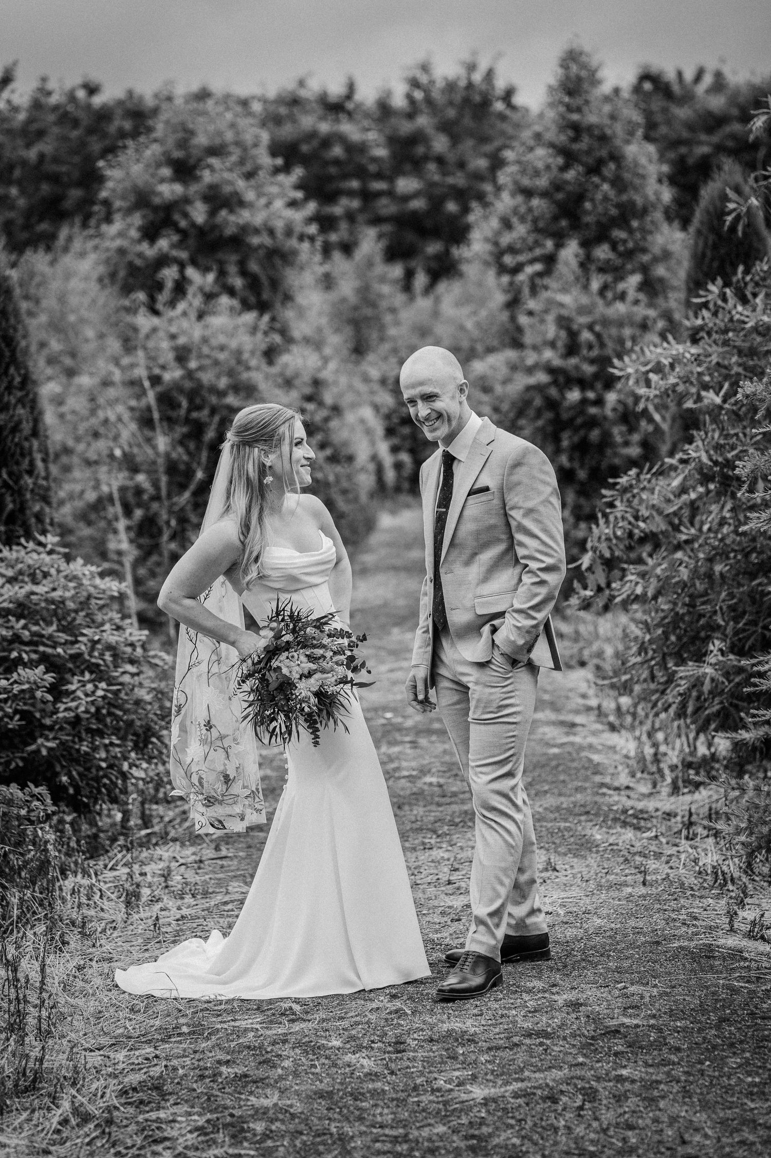 Black and white photo of a smiling bride and groom standing together in a lush garden on their wedding day.