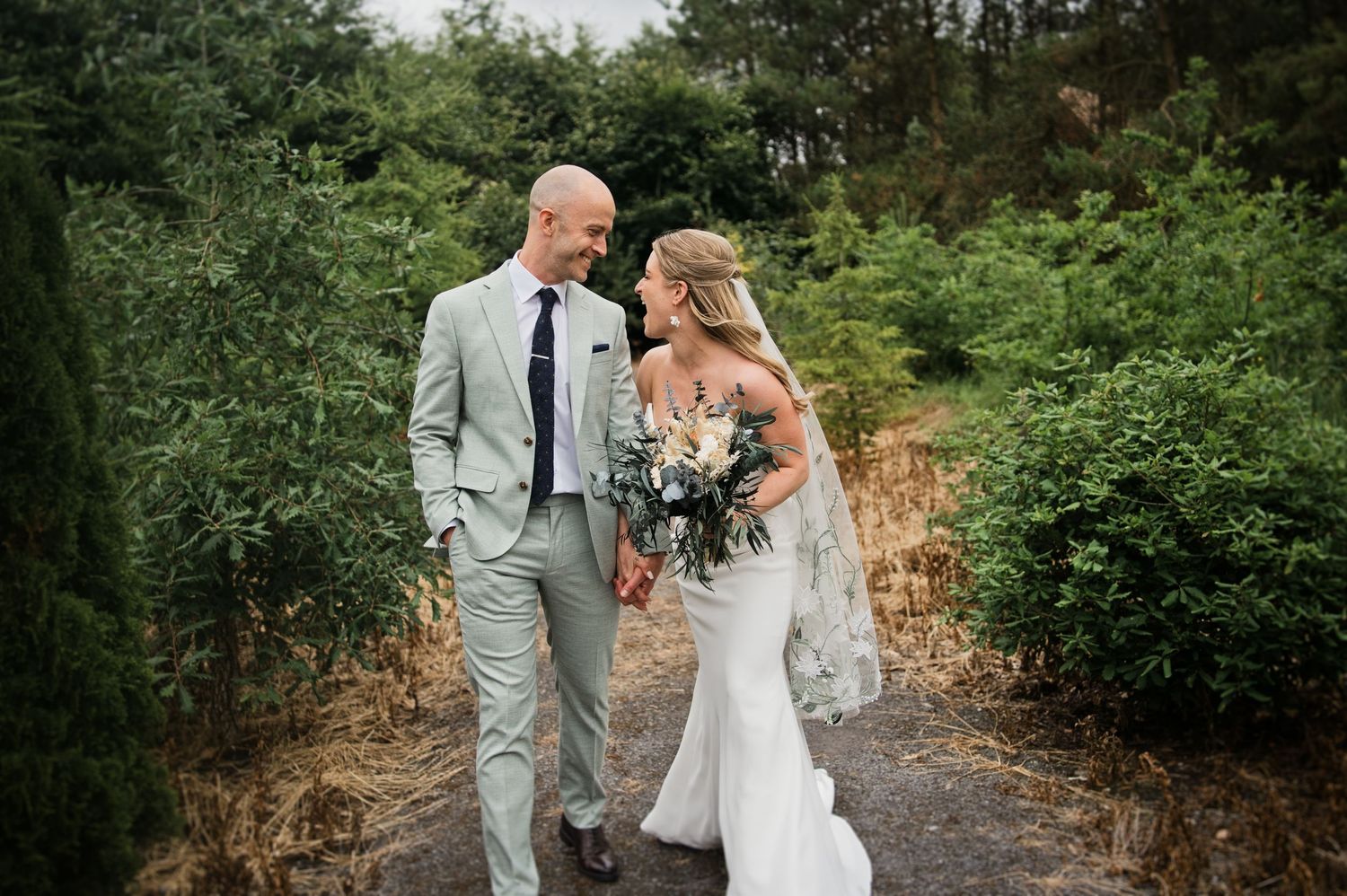 Bride and groom share a loving glance while strolling through lush greenery together.
