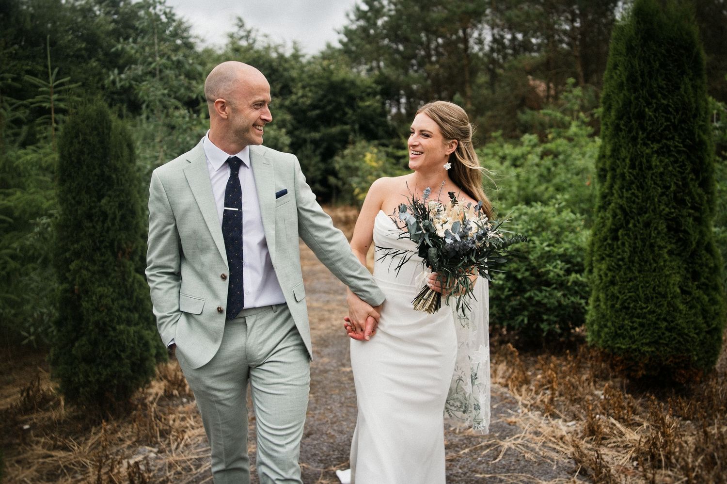 Smiling bride and groom walk hand in hand through a garden path, bride carrying a elegant bouquet.