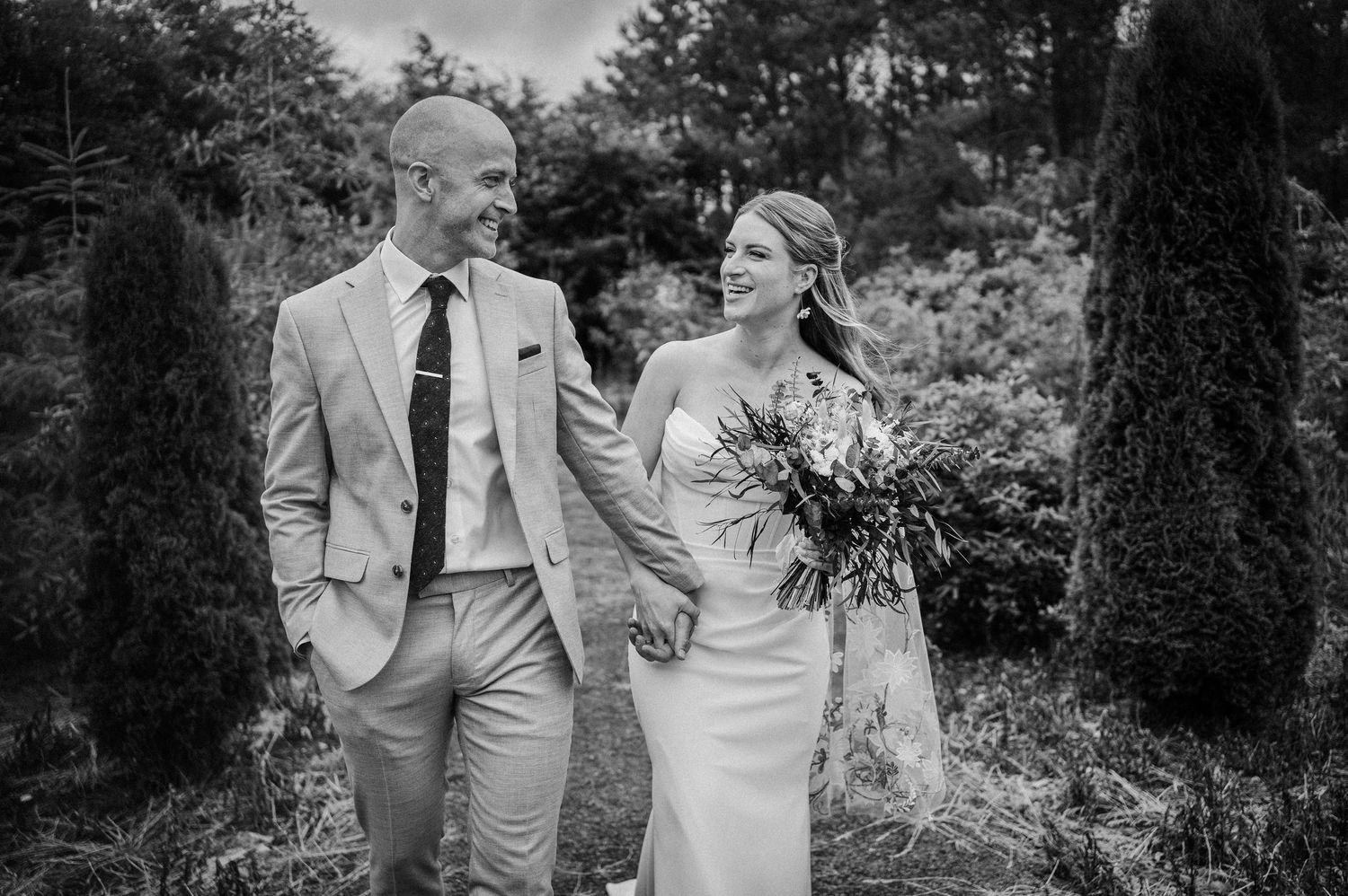 Black and white photo of a smiling bride and groom walking together outdoors, bride holding a bouquet.