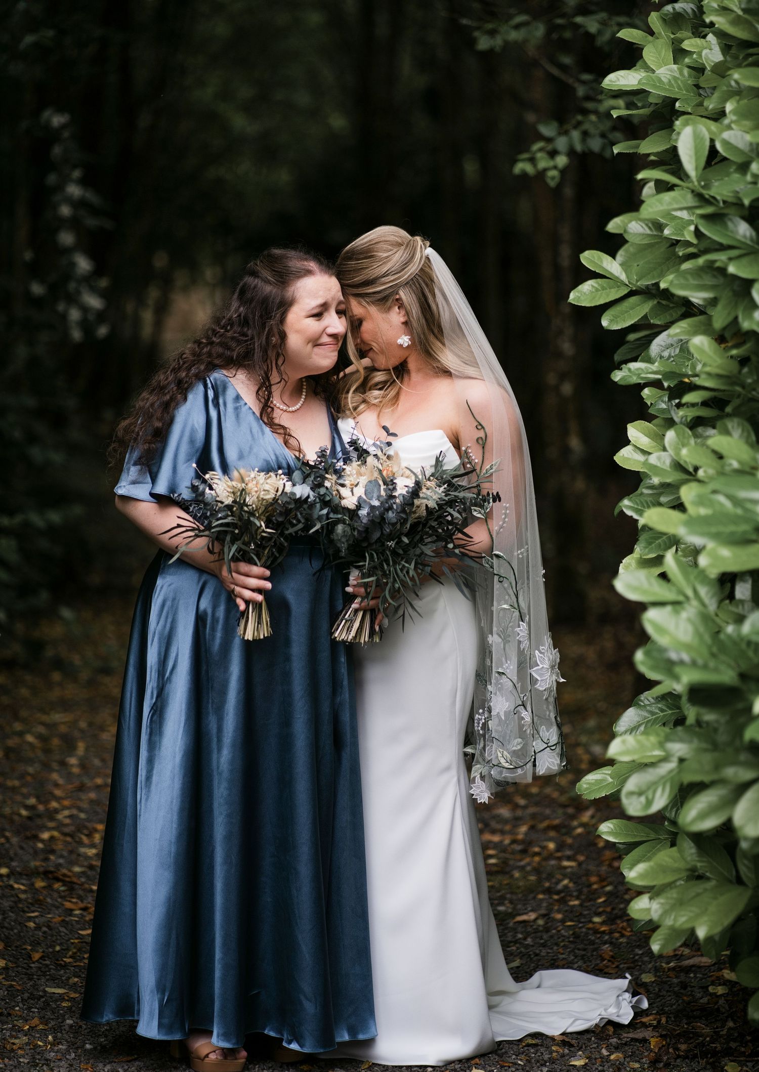 Bride in white gown and bridesmaid in blue dress share a tender moment outdoors holding bouquets.