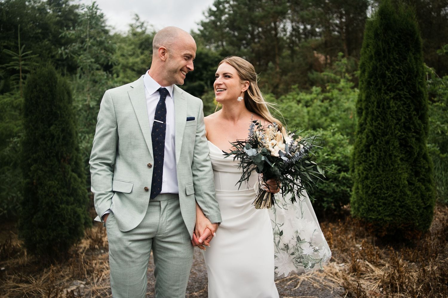 Smiling bride and groom hold hands outdoors among evergreen trees, bride holding a lush bouquet.