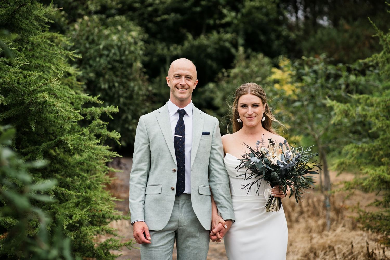 Bride and groom holding hands smiling in a lush garden setting, groom in mint suit, bride holding a bouquet.