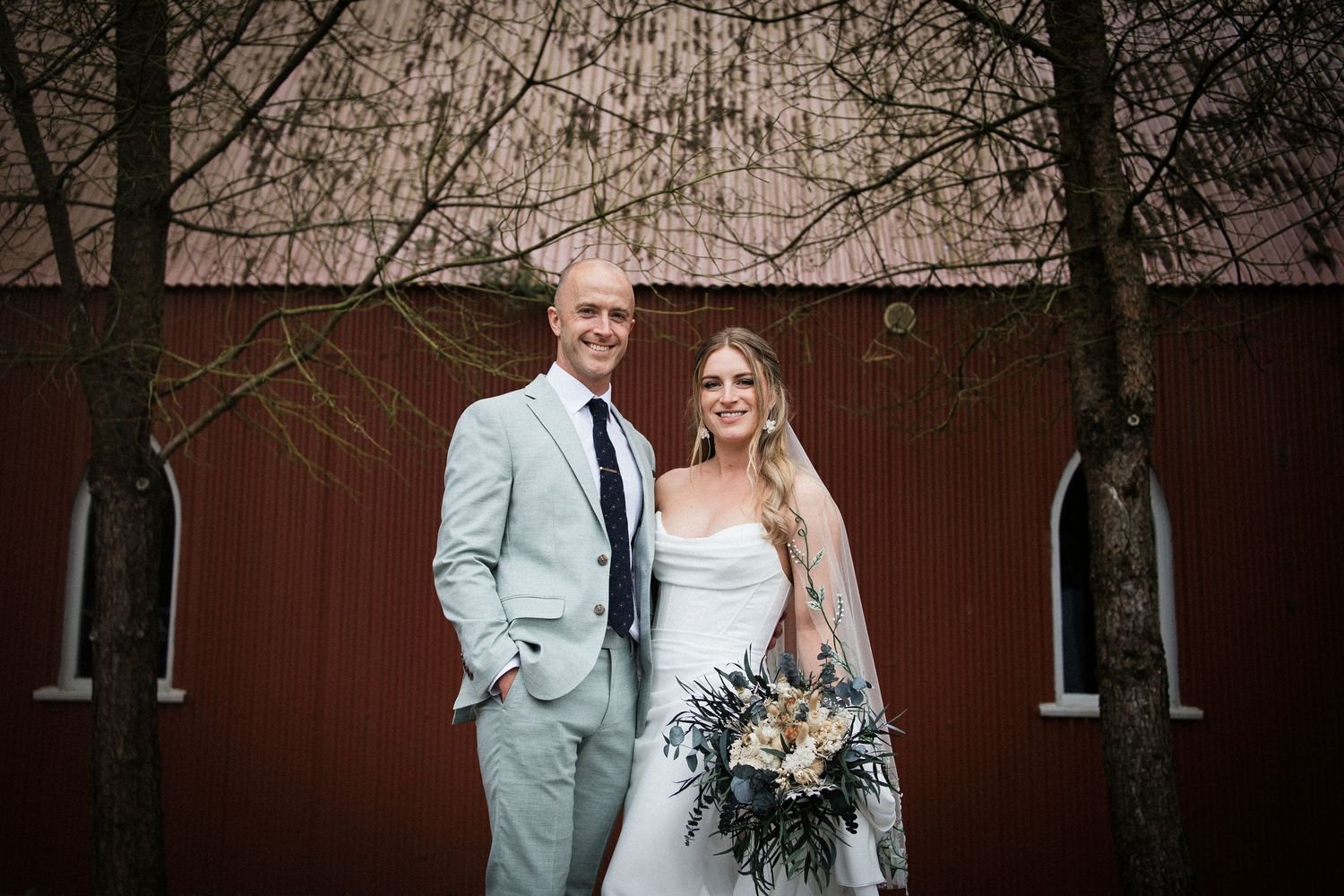 A smiling bride and groom pose together in front of a red barn on their wedding day.