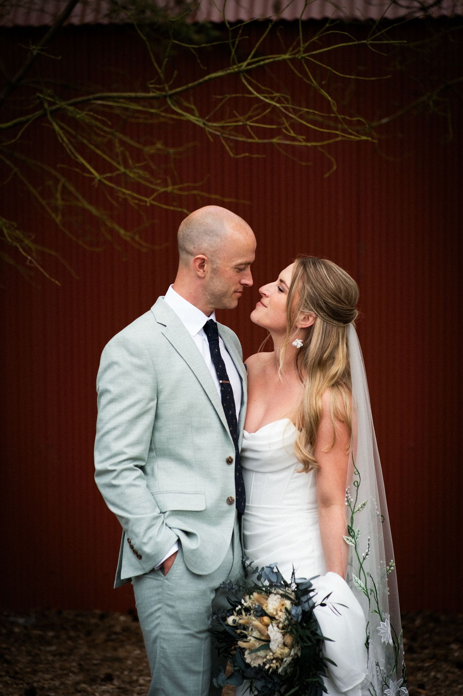 Bride and groom share a romantic moment in front of a red barn, bride holding a bouquet in a strapless gown with veil.