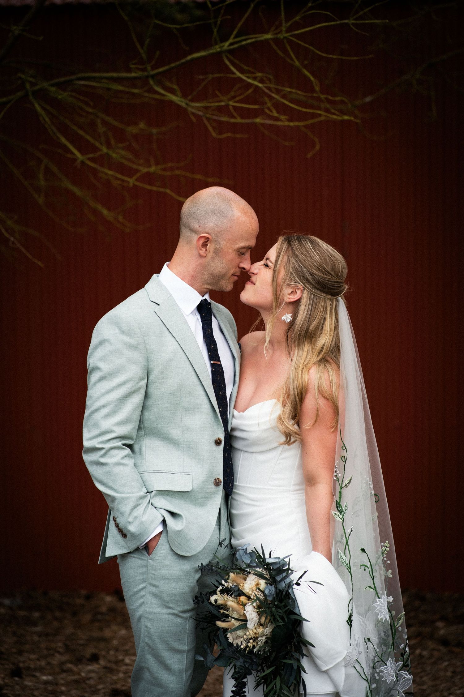 A bride and groom share a romantic almost-kiss moment in front of a rustic red barn.