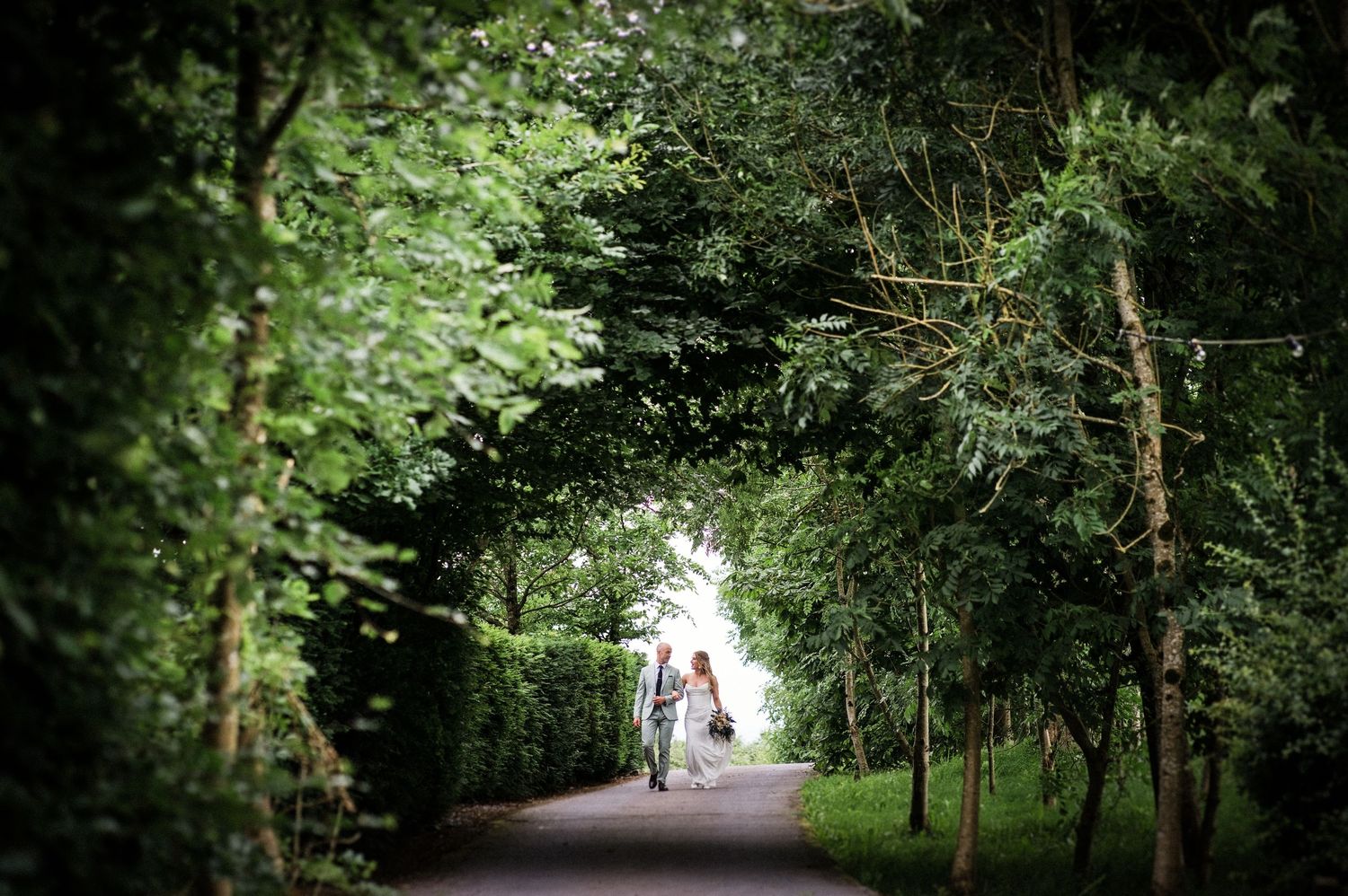 Bride and groom walk hand in hand down a tree-lined path surrounded by lush green foliage.