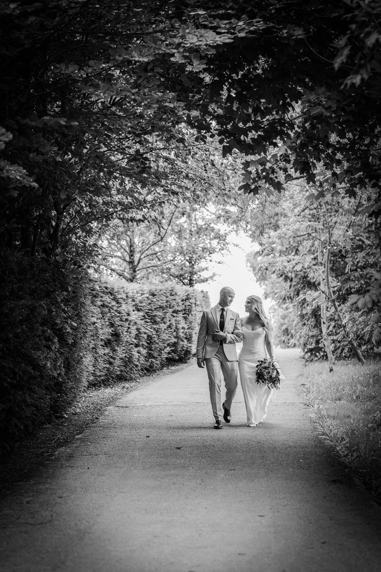 Black and white photo of bride and groom walking hand in hand along a tree-lined path on their wedding day.