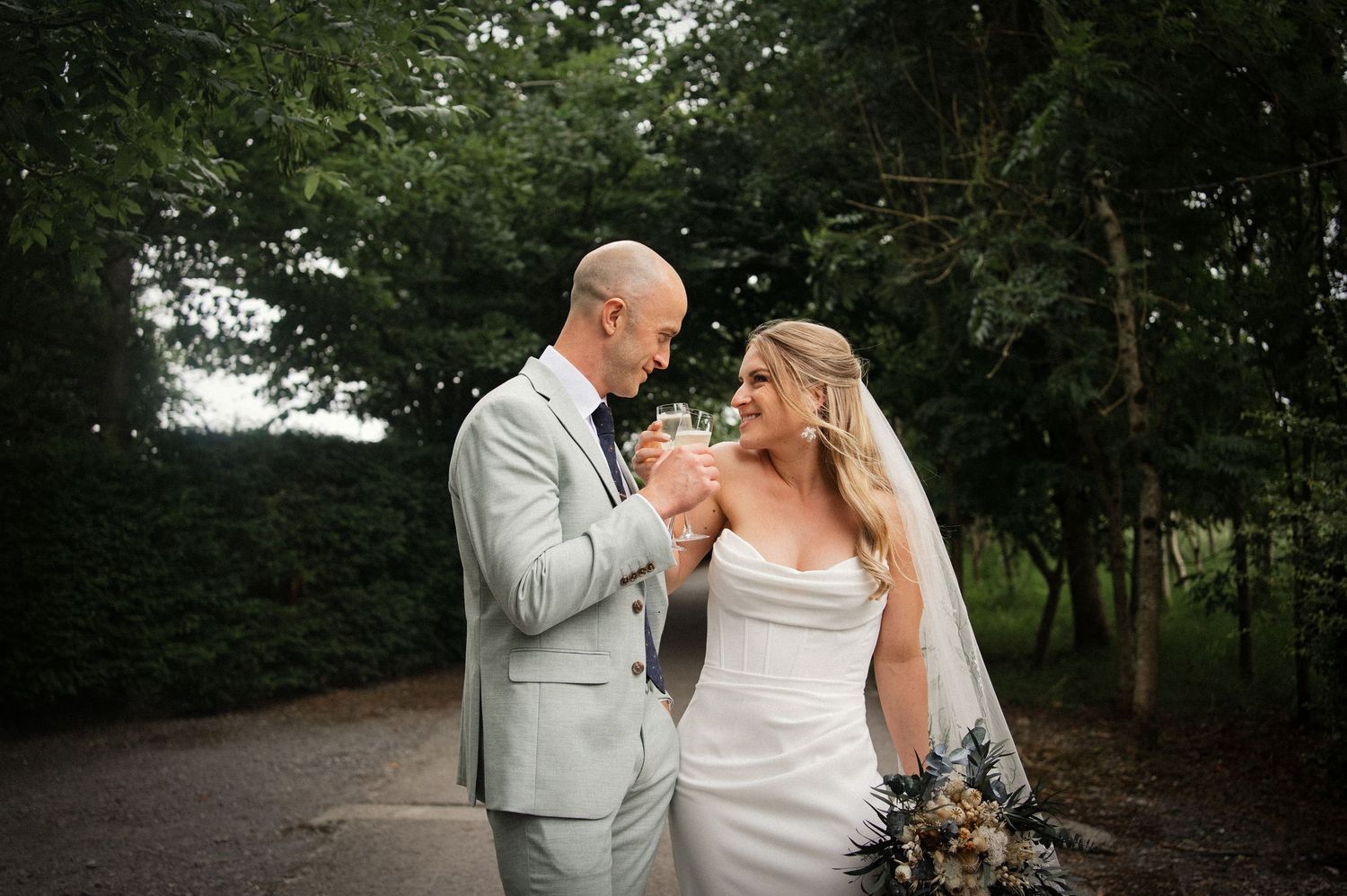 Bride and groom toasting with champagne glasses outdoors surrounded by lush green trees on their wedding day.