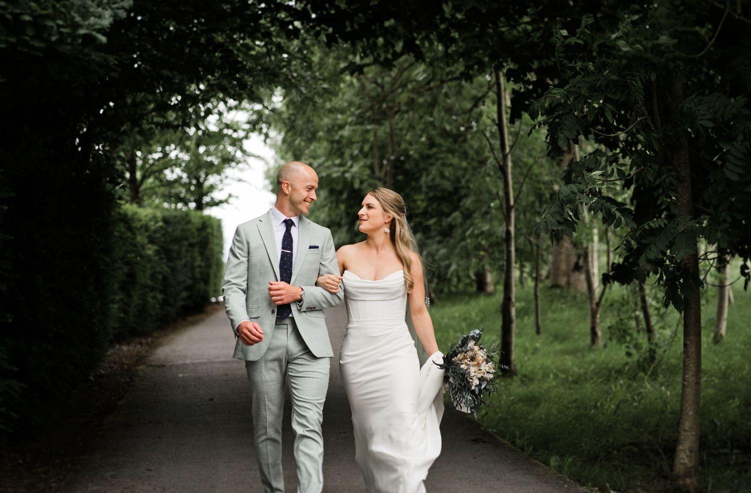 A bride and groom walk arm in arm down a tree-lined path, smiling at each other on their wedding day.
