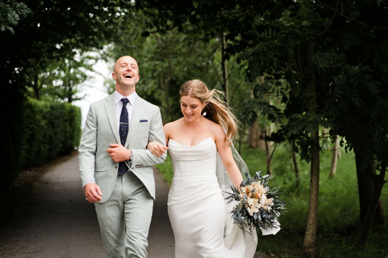 A laughing bride and groom walk hand-in-hand along a tree-lined path on their wedding day.