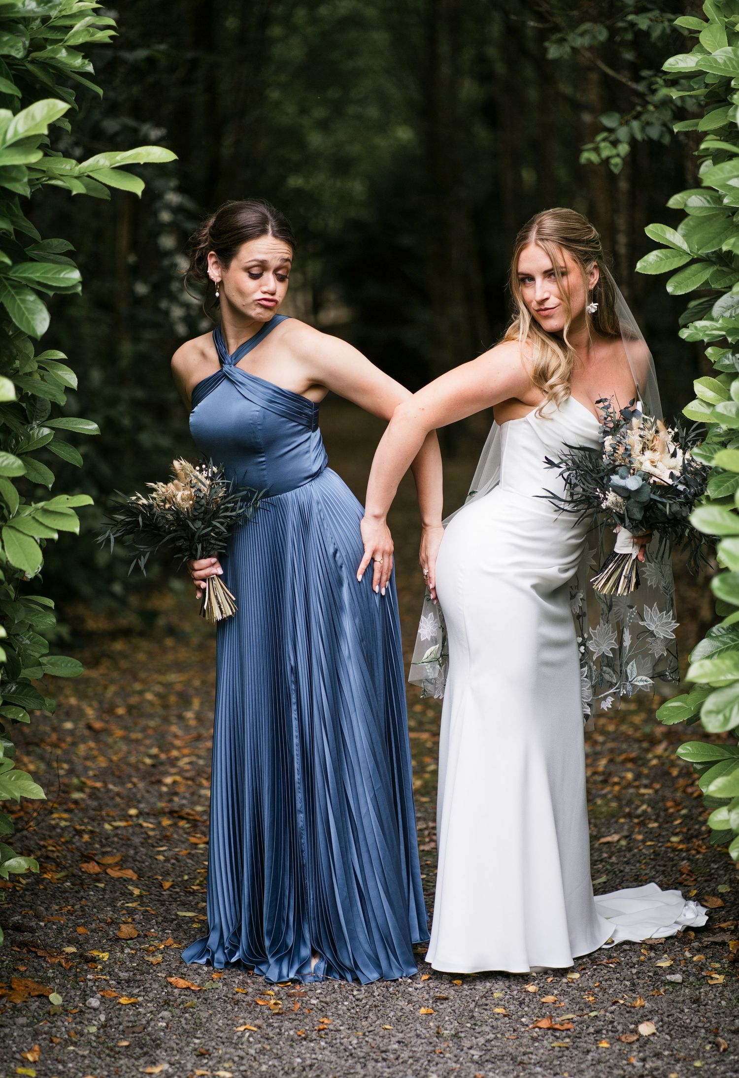 Bride in white gown and bridesmaid in blue pleated dress pose together on a garden path holding bouquets.
