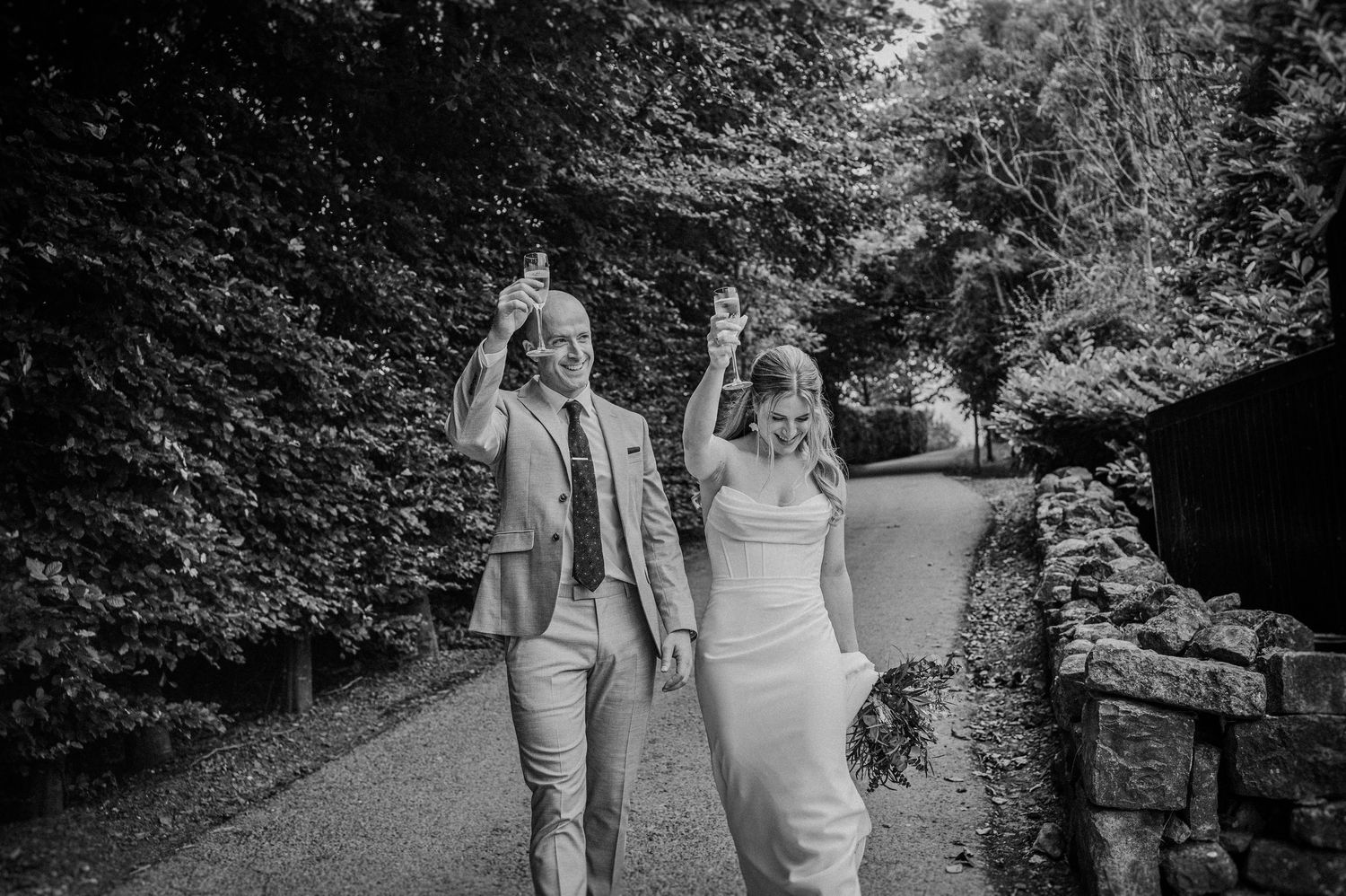 Happy couple walks down a path raising glasses in celebration on their wedding day.