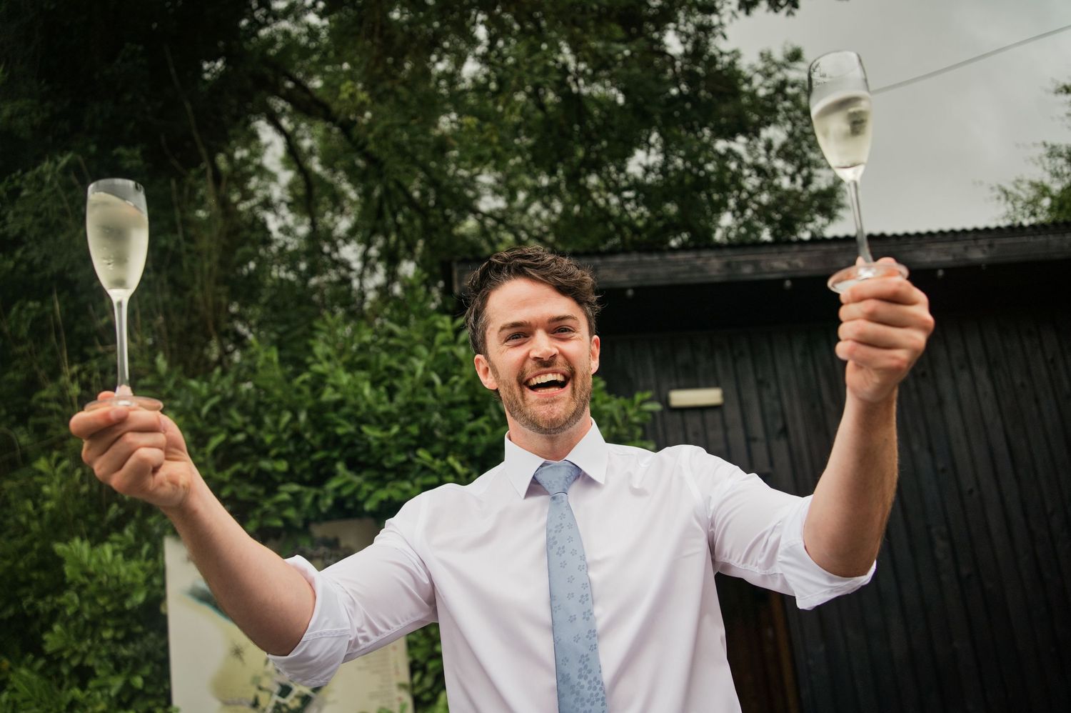 A cheerful man in a white shirt and tie raises two champagne flutes outdoors at a wedding celebration.