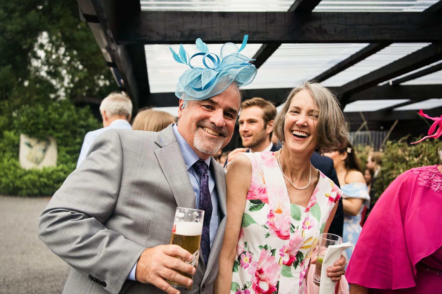A smiling couple in elegant attire enjoying drinks at an outdoor wedding reception, woman wearing a blue fascinator hat.