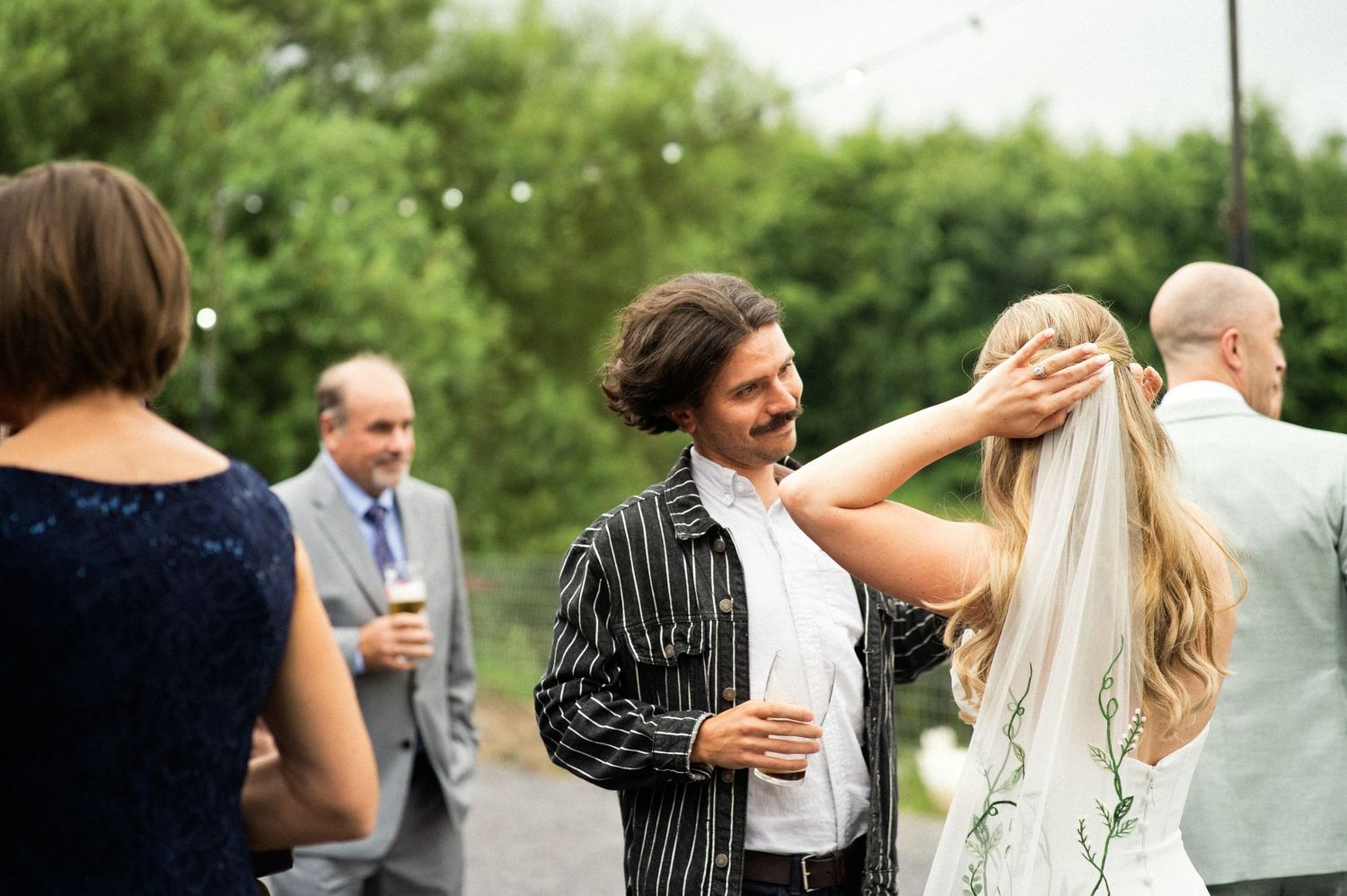 Wedding guest adjusts bride's veil during outdoor wedding reception with string lights.