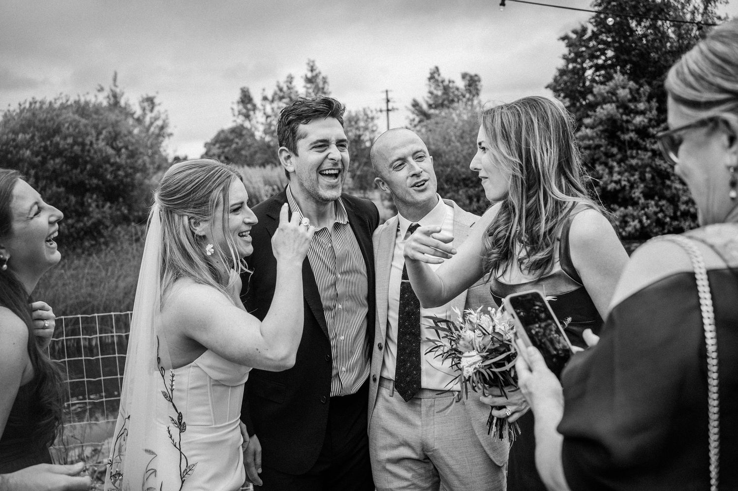 Black and white wedding photo of bride and groom laughing joyfully with guests outdoors.
