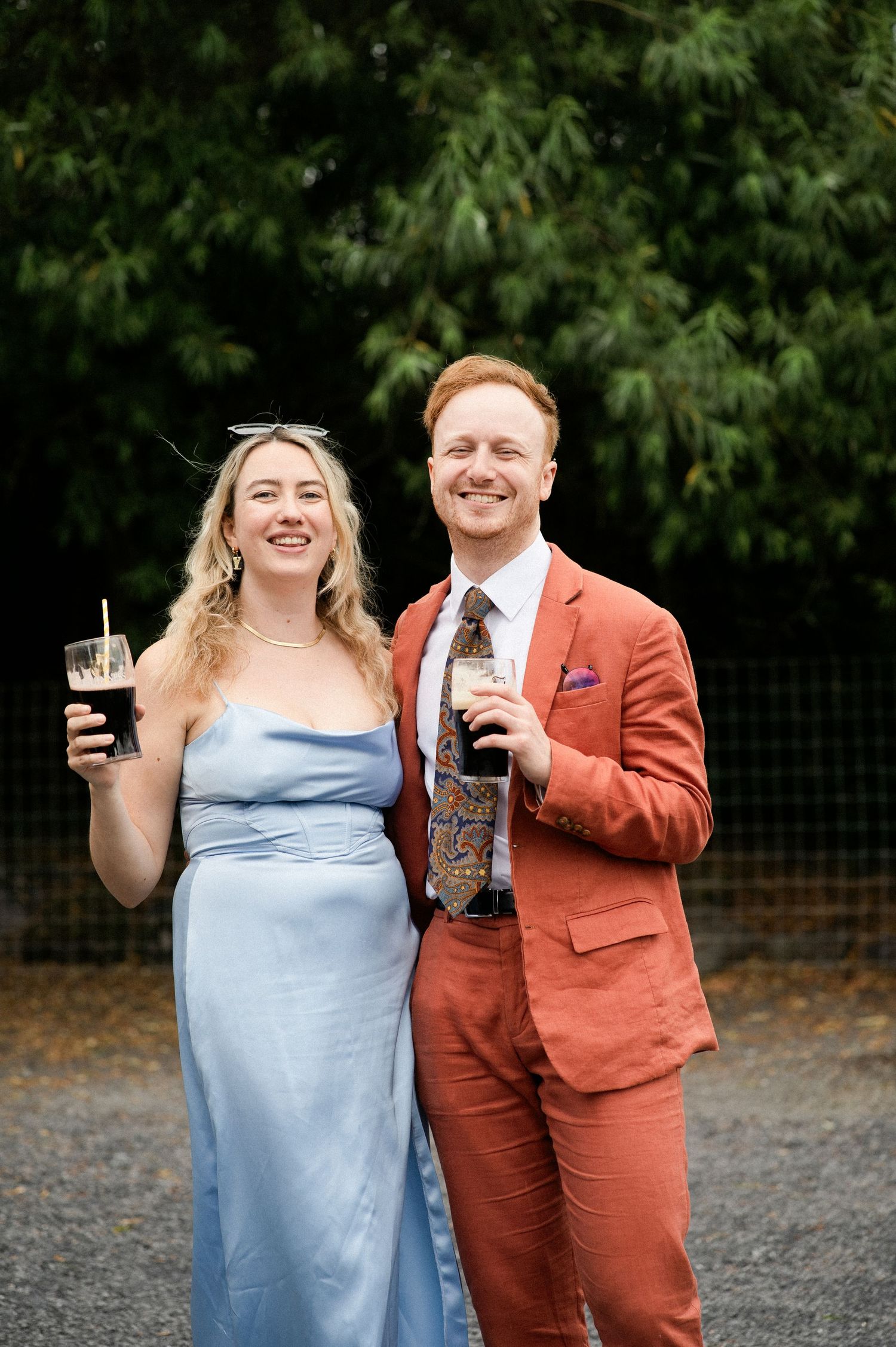 A smiling couple in elegant wedding guest attire hold drinks outdoors, the man in a rust suit and woman in a blue gown.