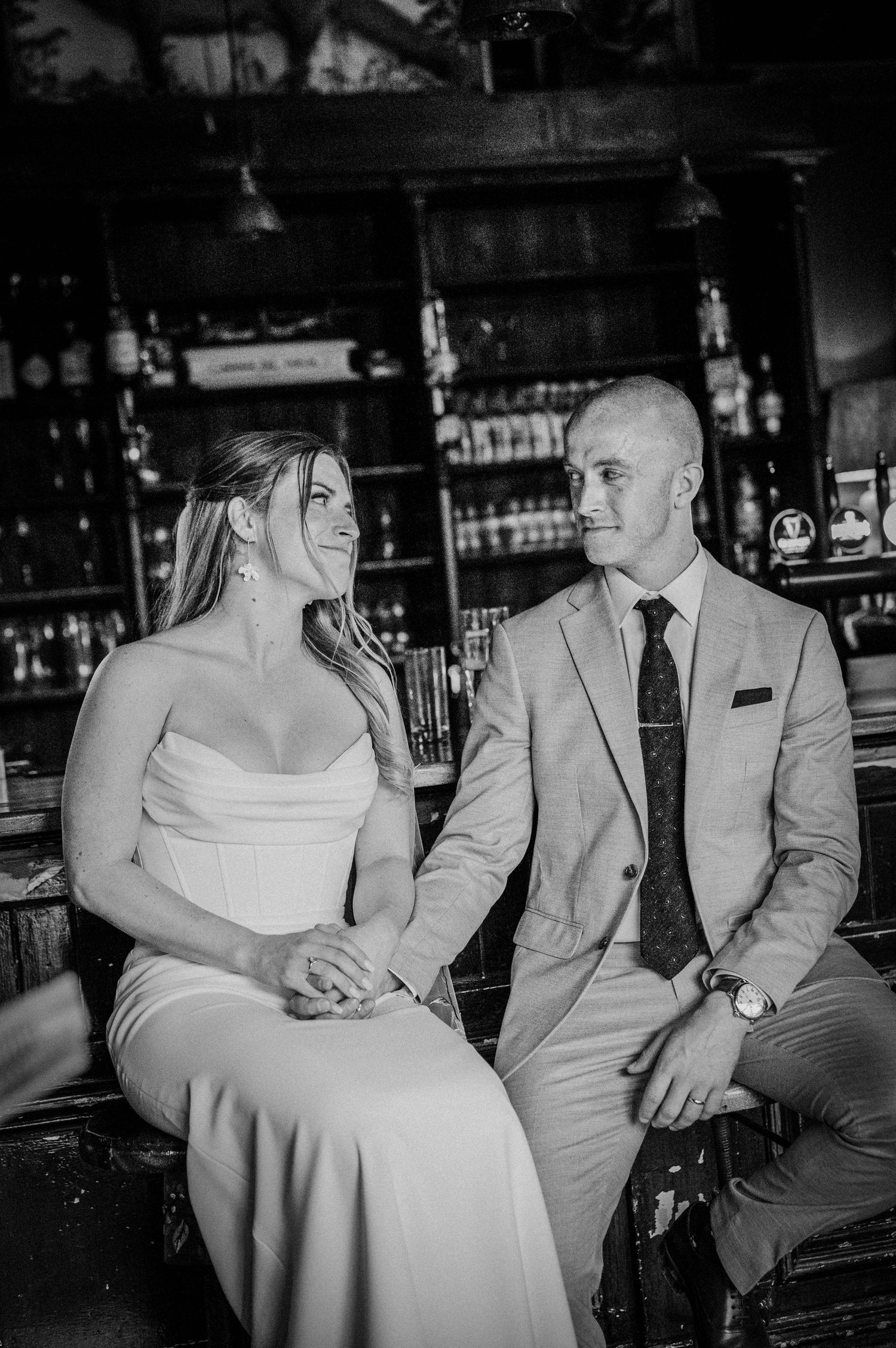 Black and white photo of bride and groom sitting together at a bar, holding hands and smiling at each other.