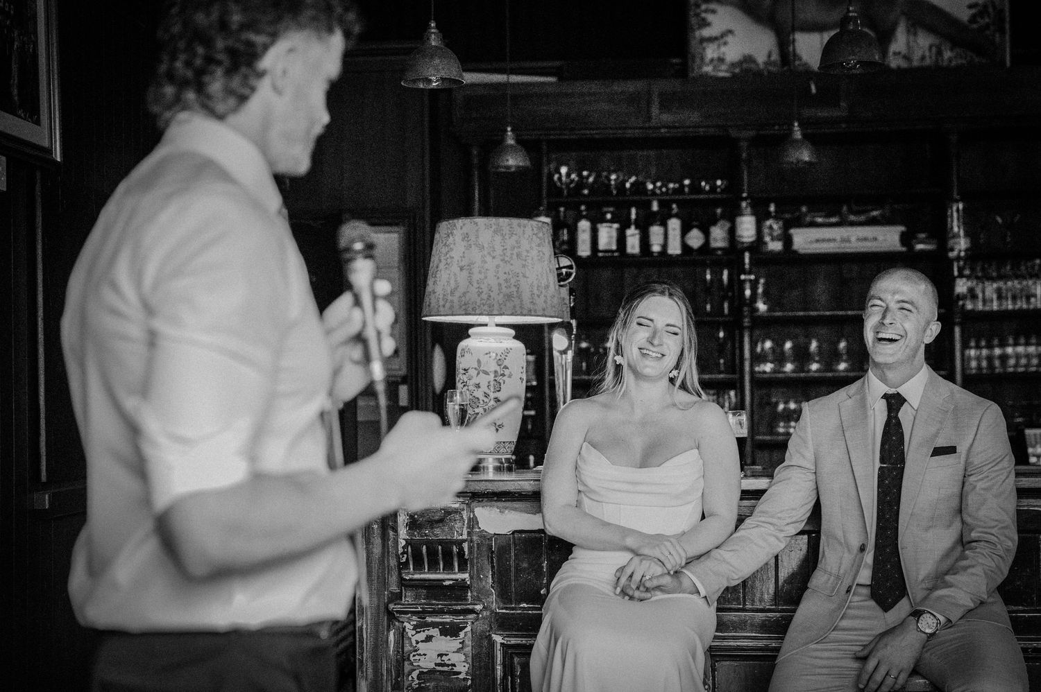A black and white photo of a guitarist performing for a laughing bride and groom seated at a bar.