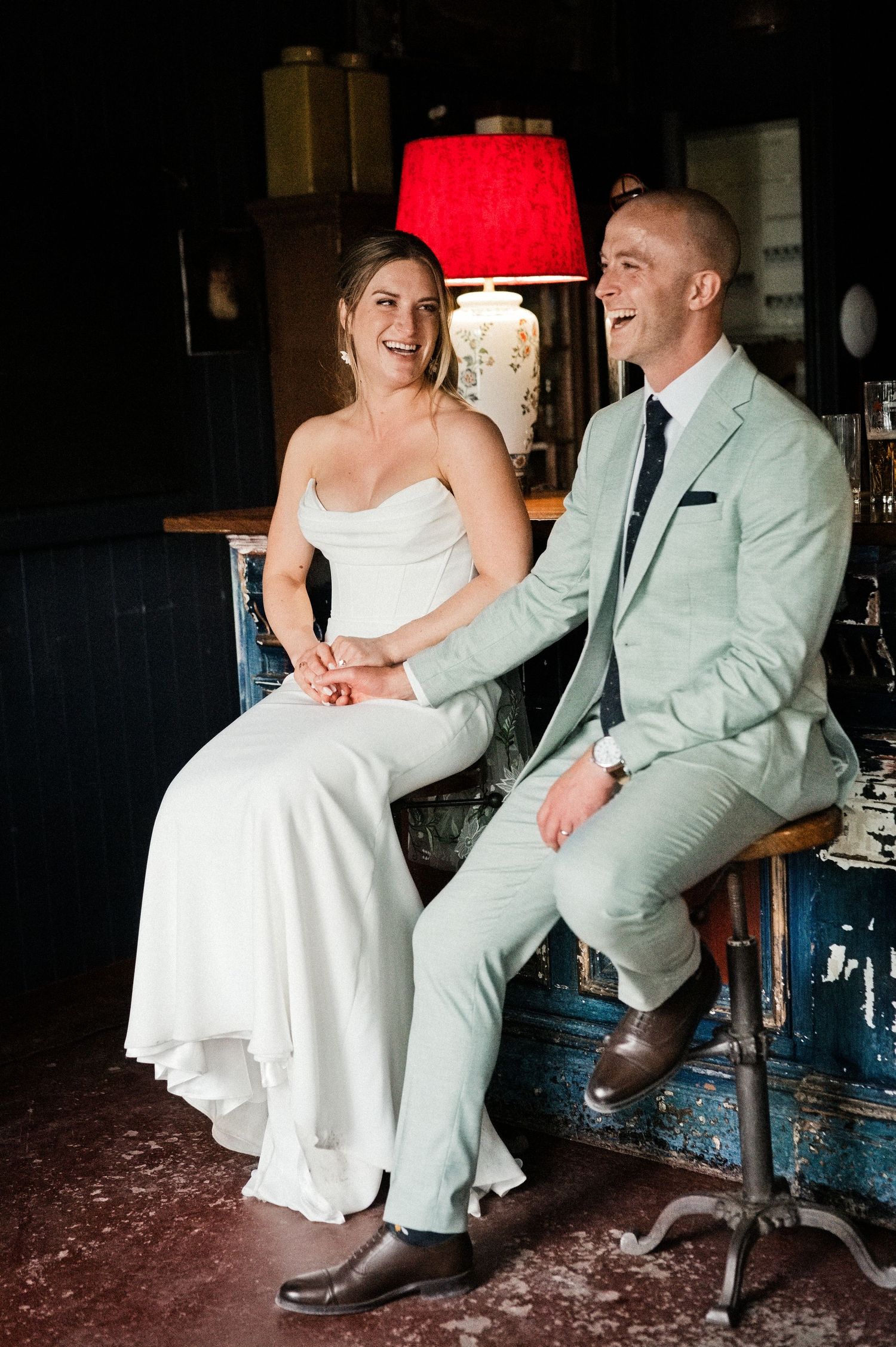 Bride in white gown and groom in mint suit laughing together while seated at a bar during their wedding reception.