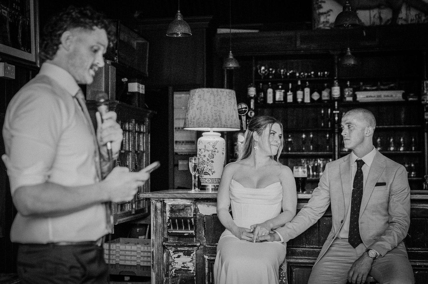 Man reads speech to bride and groom seated at bar in elegant black and white wedding photo.
