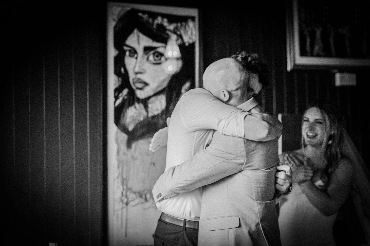 Emotional wedding moment as two people share a warm hug while bride laughs joyfully nearby.