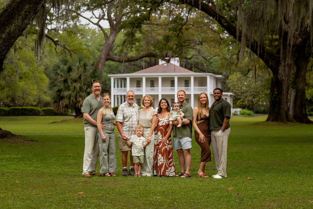 Family portrait of nine people posing on green lawn before white Southern plantation house framed by oak trees.