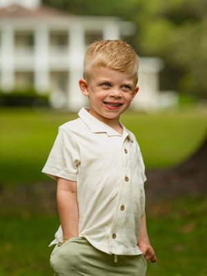 Smiling blonde boy in cream button-up shirt and green pants posing outdoors with white house in background.