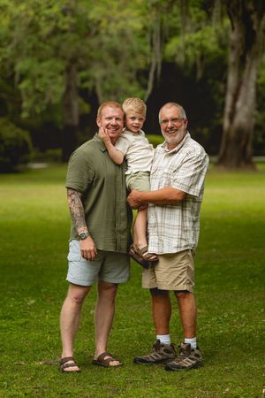 Three generations of redheaded family posing together outdoors on green grass surrounded by trees.