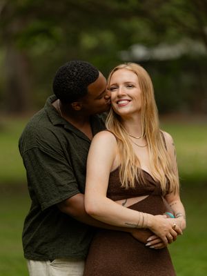 Couple embracing outdoors, man kisses smiling blonde woman in brown dress.