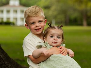 Young boy hugging baby girl outdoors near a white house, siblings posing for a family portrait on green lawn.