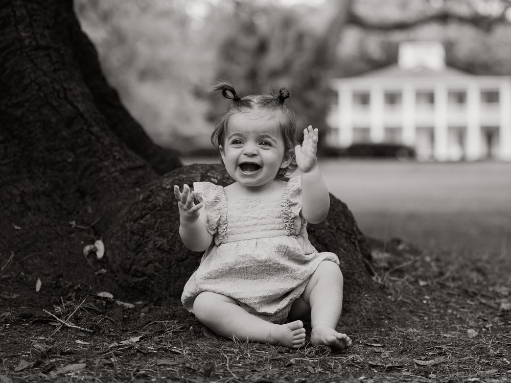 Black and white photo of a laughing baby girl sitting against a tree trunk on grass, hands raised, with a white house behind.