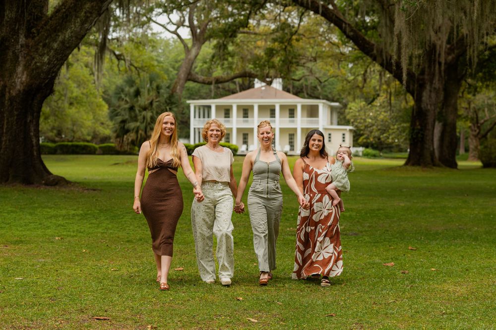 Four women walk together on green lawn before a grand white Southern plantation home with large trees.