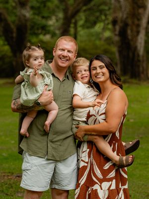 Happy family of four posing outdoors, parents holding two young children, smiling in a lush green park setting.