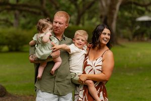 Family of four laughing together outdoors, dad holding baby girl, mom holding toddler son in a green park.