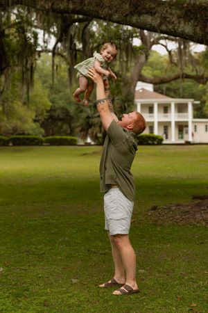 Father joyfully lifting smiling baby in green outfit above his head on lush lawn near white Southern mansion.